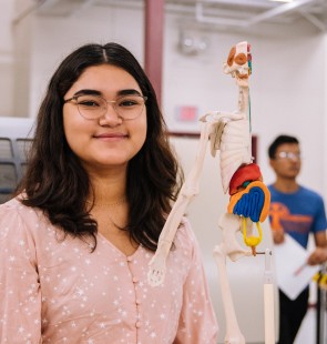 Young woman standing next to anatomical sculpture she is wearing glasses and a pink blouse.