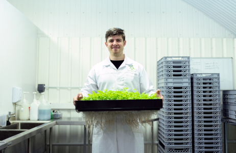 Man smiling, wearing a lab coat and carrying a bin of plants.