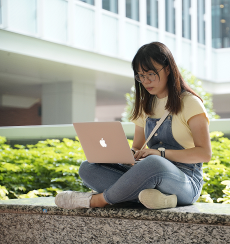 Young woman wearing glasses, sitting with her legs crossed and accessing test prep materials on her laptop. 